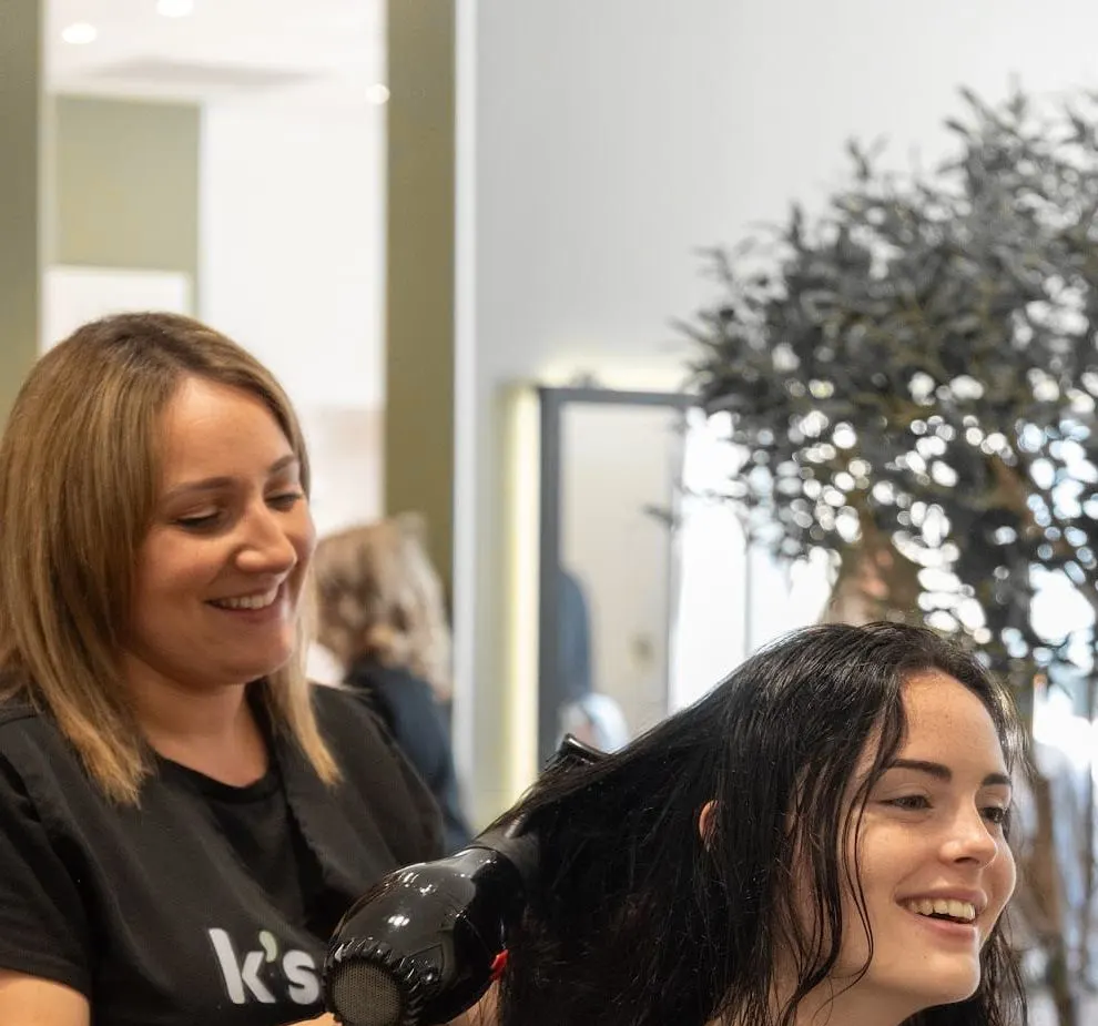 Une coiffeuse sèche en souriant les cheveux d’une cliente, qui rit.